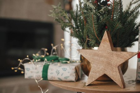 Foto de Elegante regalo de Navidad envuelto, estrella de madera y ramas de abeto en la mesa contra la chimenea en la habitación escandinava festiva. Feliz Navidad y Felices Fiestas! Imagen atmosférica - Imagen libre de derechos