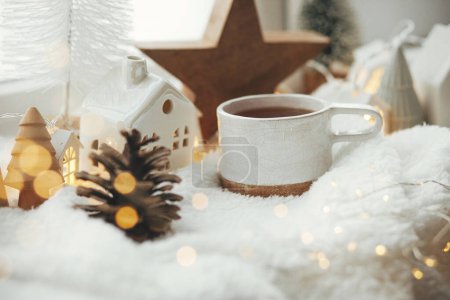 Foto de Acogedor invierno naturaleza muerta. Elegante taza de té con decoración moderna de Navidad, cono de pino, estrella de madera y árbol, luz dorada sobre una suave manta caliente en el alféizar de la ventana. Feliz Navidad y Felices Fiestas - Imagen libre de derechos