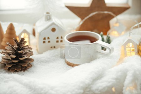 Photo for Winter hygge. Stylish cup of tea with modern cute christmas houses, pine cone, wooden star and tree, golden lights on soft warm blanket on windowsill. Christmas cozy still life. Merry Christmas! - Royalty Free Image