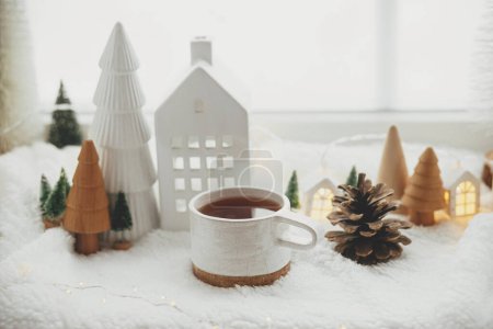 Photo for Stylish cup of tea with modern christmas houses, pine cone, wooden star and tree decor, golden lights on warm blanket on windowsill. Cozy Christmas, scandinavian style. Christmas background - Royalty Free Image
