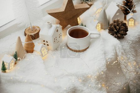 Foto de Hygge de invierno. Elegante taza de té con modernas casas de Navidad lindas, cono de pino, estrella de madera y árbol, luces doradas sobre una suave manta caliente en el alféizar de la ventana. Bodegón acogedor de Navidad. Feliz Navidad.! - Imagen libre de derechos