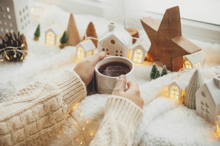 Photo for Winter hygge. Hands holding stylish cup of tea with modern cute christmas houses, pine cone, wooden star and tree, golden lights on soft warm blanket. Cozy Christmas. Happy Holidays! - Royalty Free Image