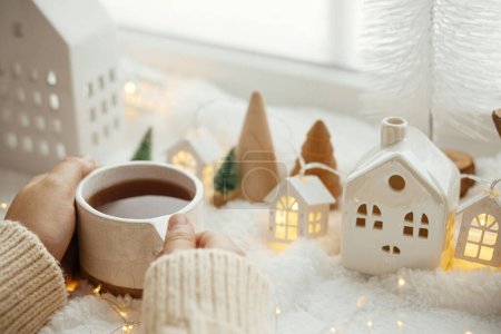 Téléchargez les photos : Un hiver douillet. Mains en pull tenant une tasse de thé élégante avec décoration de Noël moderne, cône de pin, étoile et arbre en bois, lumières dorées sur la couverture chaude douce sur le rebord de la fenêtre - en image libre de droit