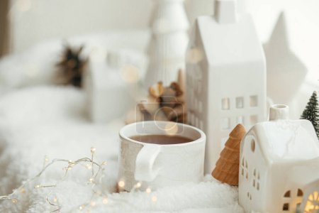 Foto de Acogedor invierno naturaleza muerta. Elegante taza de té con decoración moderna de Navidad, cono de pino, estrella de madera y árbol, luces doradas sobre una suave manta caliente en el alféizar de la ventana. Feliz Navidad y Felices Fiestas - Imagen libre de derechos