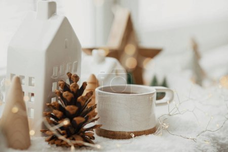 Foto de Elegante taza de té con casas de Navidad modernas, cono de pino, estrella de madera y decoración de árboles, luces doradas en una manta caliente en el alféizar de la ventana. Acogedora Navidad, estilo escandinavo. Fondo de Navidad - Imagen libre de derechos