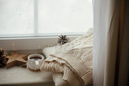 Foto de Elegante taza de té con acogedor suéter de punto, cono de pino, estrella de madera y luces doradas en el alféizar de la ventana en la habitación escandinava festiva. Acogedor invierno naturaleza muerta. Hygge otoñal - Imagen libre de derechos
