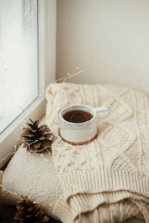 Foto de Acogedor invierno naturaleza muerta. Elegante taza de té con acogedor suéter de punto, cono de pino, estrella de madera y luces doradas en el alféizar de la ventana en la habitación escandinava festiva. Vacaciones de otoño y Navidad - Imagen libre de derechos