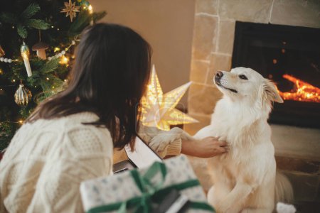 Photo for Happy woman holding stylish christmas gifts with credit card and caressing cute white dog in festive decorated christmas room with lights. Christmas holidays and pet - Royalty Free Image