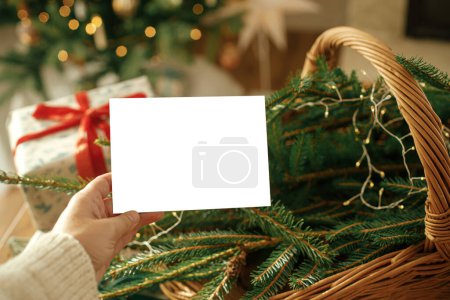 Téléchargez les photos : Main tenant carte de vœux vide sur fond de cadeaux de Noël enveloppés élégants, branches de sapin dans un panier rustique et arbre de fête avec des lumières dorées. Carte de Noël maquillée. Espace pour le texte - en image libre de droit