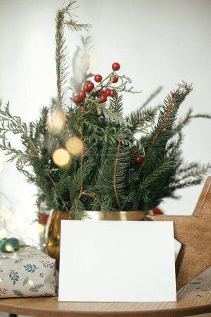 Foto de La tarjeta de Navidad se burla. Tarjeta de felicitación vacía, elegante regalo de Navidad envuelto, estrella rústica y ramas de abeto con luces en la mesa de madera. Espacio para el texto. Plantilla de postal de felicitaciones de temporada - Imagen libre de derechos