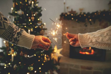 Foto de ¡Feliz Año Nuevo! Bengalas ardientes en las manos en el fondo de la chimenea del país moderno y el árbol de Navidad con luces doradas. Fuegos artificiales brillando en las manos, pareja celebrando en habitación decorada festiva - Imagen libre de derechos