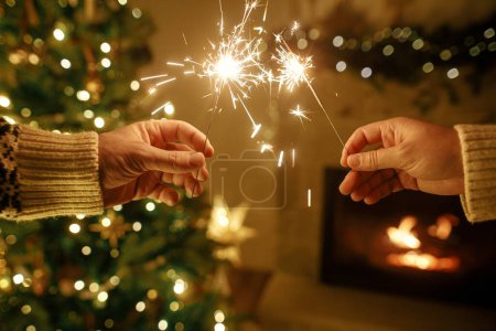 Photo for Happy New Year! Burning sparklers in hands  in eve on background of modern fireplace and christmas tree with golden lights. Fireworks glowing in hands, couple celebrating in festive dark room - Royalty Free Image