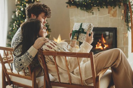 Photo for Happy couple in cozy sweaters exchanging stylish wrapped christmas gifts on background of fireplace with festive mantle and modern decorated christmas tree with lights. Merry Christmas! - Royalty Free Image