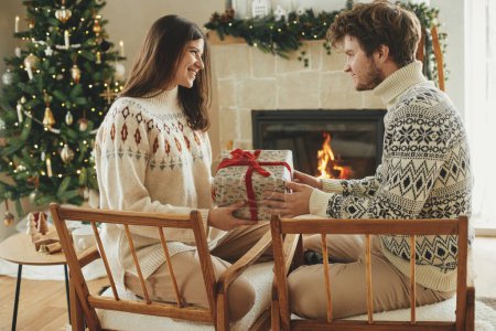 Photo for Happy young family in cozy sweaters exchanging stylish christmas gifts on background of fireplace with modern festive mantle and christmas tree with lights. Happy Holidays! - Royalty Free Image