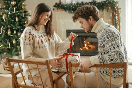 Photo for Happy couple in cozy sweaters opening stylish wrapped christmas gifts on background of fireplace with festive mantle and modern decorated christmas tree with lights. Merry Christmas! - Royalty Free Image