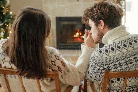 Téléchargez les photos : Couple élégant en pulls confortables embrassant et relaxant à la cheminée avec manteau festif sur fond de sapin de Noël décoré élégant avec des lumières. Joyeux jeune famille profitant des vacances d'hiver - en image libre de droit