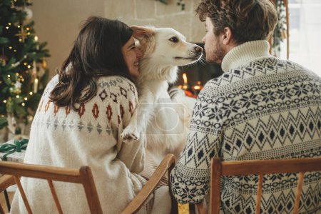 Photo for Happy young family in cozy sweaters hugging with cute dog and relaxing on background of fireplace and modern christmas tree. Merry Christmas! Pets and winter holidays - Royalty Free Image