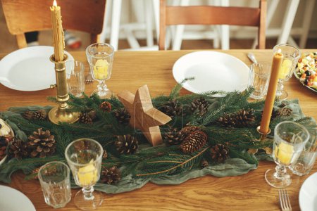 Photo for Christmas rustic table setting. Stylish table runner with fir branches and pine cones, candles, cheese appetizers and starters, glasses and cutlery. Holiday dinner arrangement. Happy new year! - Royalty Free Image