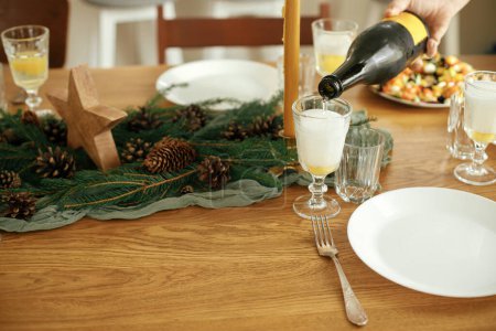 Photo for Christmas festive dinner. Hands pouring champagne into glasses on wooden table with fir branches and pine cones decor, appetizers, glasses and cutlery. Happy new year! Holiday arrangement - Royalty Free Image