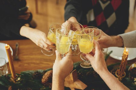 Téléchargez les photos : Amis et famille célébrant et grillant au champagne. Mains avec verres à cocktail mimosa en cliquant sur le fond de table de Noël élégant avec branches de sapin, cônes de pin et bougies - en image libre de droit