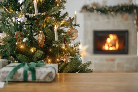 Téléchargez les photos : Joyeux Noël ! Élégant arbre décoré de Noël, lumières dorées, cadeau sur la table et cheminée en feu. Veille de Noël atmosphérique à la cheminée, espace pour le texte - en image libre de droit