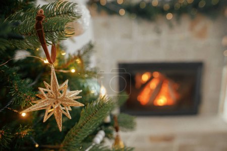 Photo for Merry Christmas! Stylish christmas golden star on tree close up against burning fireplace. Beautiful decorated christmas tree with vintage baubles and ribbons. Christmas background - Royalty Free Image