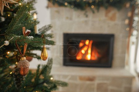 Photo for Stylish christmas acorn bauble on tree close up against burning fireplace. Beautiful decorated christmas tree with vintage ornaments, ribbons and lights. Christmas background. Merry Christmas! - Royalty Free Image