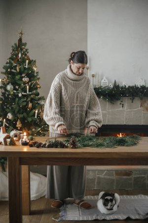 Foto de Mujer en suéter acogedor haciendo corona rústica de Navidad en la mesa de madera con gato lindo en la sala de estar festiva con chimenea. Preparativos para las vacaciones de invierno, tiempo atmosférico - Imagen libre de derechos