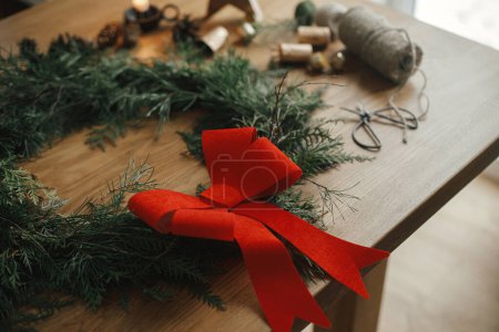 Téléchargez les photos : Élégante couronne rustique de Noël avec arc rouge, branches de cèdre, cloches vintage, cônes de pin, ficelle, ciseaux sur table en bois. Joyeux Noël. Préparation des vacances d'hiver dans la salle atmosphérique - en image libre de droit
