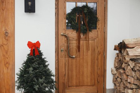 Foto de Elegante corona rústica de Navidad con campanas vintage y cinta en puertas de madera y abeto con lazo rojo en la entrada de la casa. Decoración de vacaciones de invierno de exterior de granja moderna - Imagen libre de derechos