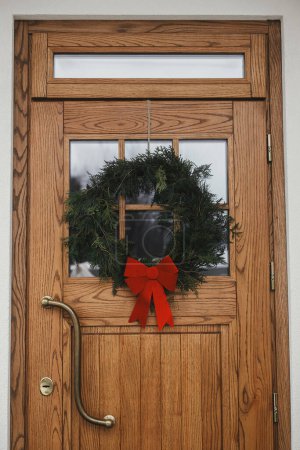 Téléchargez les photos : Couronne de Noël élégante avec arc rouge accroché sur la porte d'entrée en bois à l'entrée de la maison. Moderne décor extérieur minimal de ferme pour les vacances d'hiver. Joyeux Noël et Joyeuses fêtes - en image libre de droit