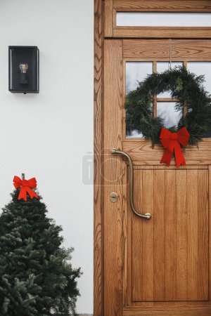 Photo for Stylish christmas rustic wreath with red bow on wooden doors and fir tree with red bow at house entrance. Winter holiday decoration of modern farmhouse exterior - Royalty Free Image