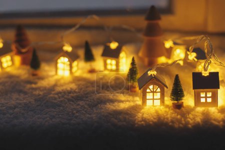 Photo for Cozy christmas miniature village. Stylish cute little glowing houses and christmas trees on soft snow blanket with lights in evening room. Atmospheric winter village still life. Merry Christmas! - Royalty Free Image