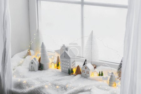 Foto de Aldea invernal en miniatura atmosférica. Pequeñas y elegantes casas de cerámica y árboles de madera de Navidad en una suave manta de nieve con luces brillantes en la habitación. Navidad moderno fondo blanco. - Imagen libre de derechos