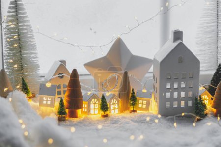 Téléchargez les photos : Village d'hiver miniature atmosphérique. Élégantes petites maisons en céramique et des arbres de Noël en bois sur la couverture de neige avec des lumières incandescentes en soirée. Noël fond moderne. Joyeuses fêtes! - en image libre de droit