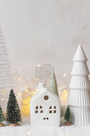 Téléchargez les photos : Confortable village miniature de Noël. Élégantes petites maisons en céramique et des arbres sur la couverture de neige avec des lumières dorées sur fond blanc. Village d'hiver atmosphérique moderne nature morte. Joyeux Noël! - en image libre de droit