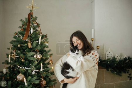 Téléchargez les photos : Femme heureuse en pull confortable étreignant chat mignon sur fond d'arbre de Noël décoré élégant dans la salle de fête. Propriétaire avec animaux et vacances d'hiver. Joyeux Noël! - en image libre de droit