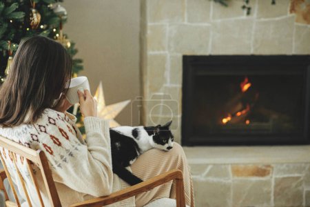 Foto de Vacaciones de invierno Hygge. Mujer en suéter acogedor sentado con gato lindo y beber té caliente en el fondo de la chimenea y elegante árbol de Navidad. Propietario con mascota relajante. Feliz Navidad.! - Imagen libre de derechos
