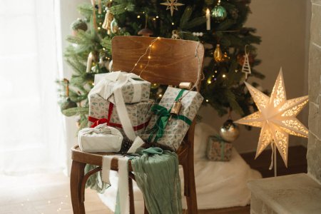 Photo for Stylish wrapped christmas gifts with ribbon on wooden old chair on background of decorated christmas tree with vintage baubles and festive lights. Merry Christmas and Happy Holidays! - Royalty Free Image