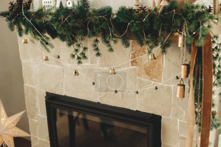 Téléchargez les photos : Manteau cheminée élégant décoré avec branches de Noël, guirlande de cloches, ornements en bois et décoration de la maison. Cheminée de Noël festive dans le salon moderne de la ferme - en image libre de droit