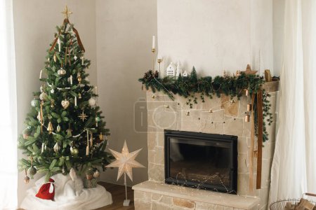 Photo for Stylish decorated fireplace mantel with christmas branches, decorations and bells on background of modern christmas tree with lights. Rustic christmas living room in modern farmhouse - Royalty Free Image