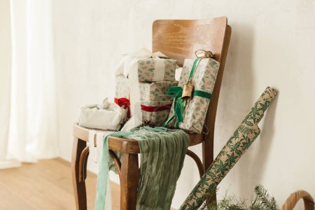 Téléchargez les photos : Joyeux Noël et Joyeuses Fêtes ! Élégants cadeaux de Noël enveloppés avec des rubans sur une vieille chaise en bois, du papier de fête et un panier en osier avec des branches d'épinette dans une pièce rurale. Cadeaux respectueux de l'environnement - en image libre de droit