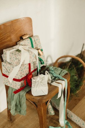 Téléchargez les photos : Joyeux Noël et Joyeuses Fêtes ! Élégants cadeaux de Noël enveloppés avec des rubans sur une vieille chaise en bois, du papier de fête et un panier en osier avec des branches d'épinette dans une pièce rurale. Cadeaux respectueux de l'environnement - en image libre de droit