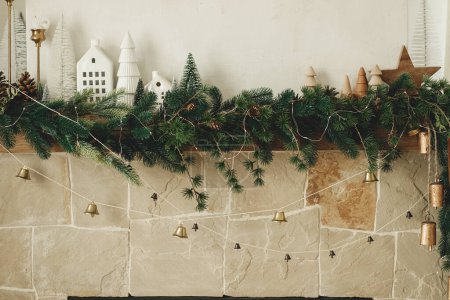 Foto de Elegante chimenea mantel decorado con ramas de Navidad, guirnalda campanas, adornos de madera y decoración de la casa. Chimenea de Navidad rústica festiva en la moderna casa de campo sala de estar - Imagen libre de derechos
