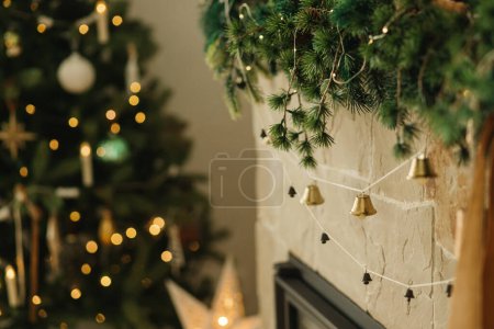 Foto de Elegante chimenea mantel decorado con ramas de Navidad, guirnalda campanas, adornos en el fondo de las luces del árbol de Navidad. Decoración festiva chimenea rústica en la moderna casa de campo sala de estar - Imagen libre de derechos