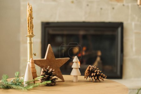Téléchargez les photos : Élégants arbres de Noël en bois, bougie, étoile, cônes de pin et branches de sapin sur la table contre la cheminée en feu. Moderne décor rustique écologique pour des vacances d'hiver dans le salon de la ferme - en image libre de droit