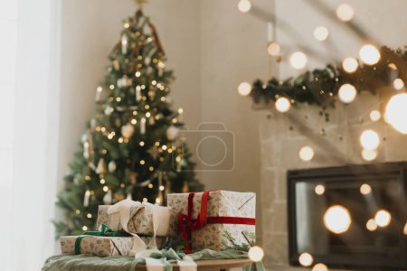 Téléchargez les photos : Cadeaux de Noël enveloppés élégants avec ruban sur la table sur fond d'arbre de Noël moderne décoré avec des boules vintage, cheminée rustique et lumières festives. Joyeux Noël et Joyeuses Fêtes! - en image libre de droit