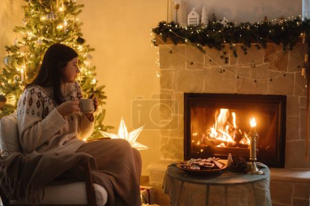 Foto de Mujer en suéter acogedor celebración taza de té y relajarse en la chimenea en el fondo del elegante árbol de Navidad con luces en la habitación festiva de la noche. Vacaciones de invierno hygge. Nochebuena - Imagen libre de derechos