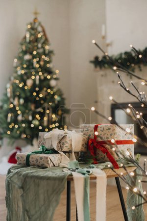 Téléchargez les photos : Joyeux Noël et Joyeuses Fêtes ! Cadeaux de Noël enveloppés élégants avec ruban sur la table sur fond d'arbre de Noël décoré avec des boules vintage, cheminée rustique et lumières festives - en image libre de droit