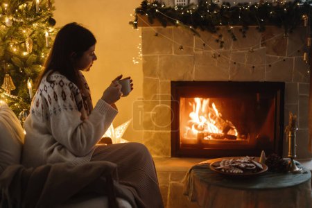 Téléchargez les photos : Femme en pull confortable tenant une tasse de thé et se réchauffant à la cheminée brûlante sur fond d'arbre de Noël élégant avec des lumières dans la salle de fête du soir. Vacances d'hiver hygge. Veille de Noël - en image libre de droit